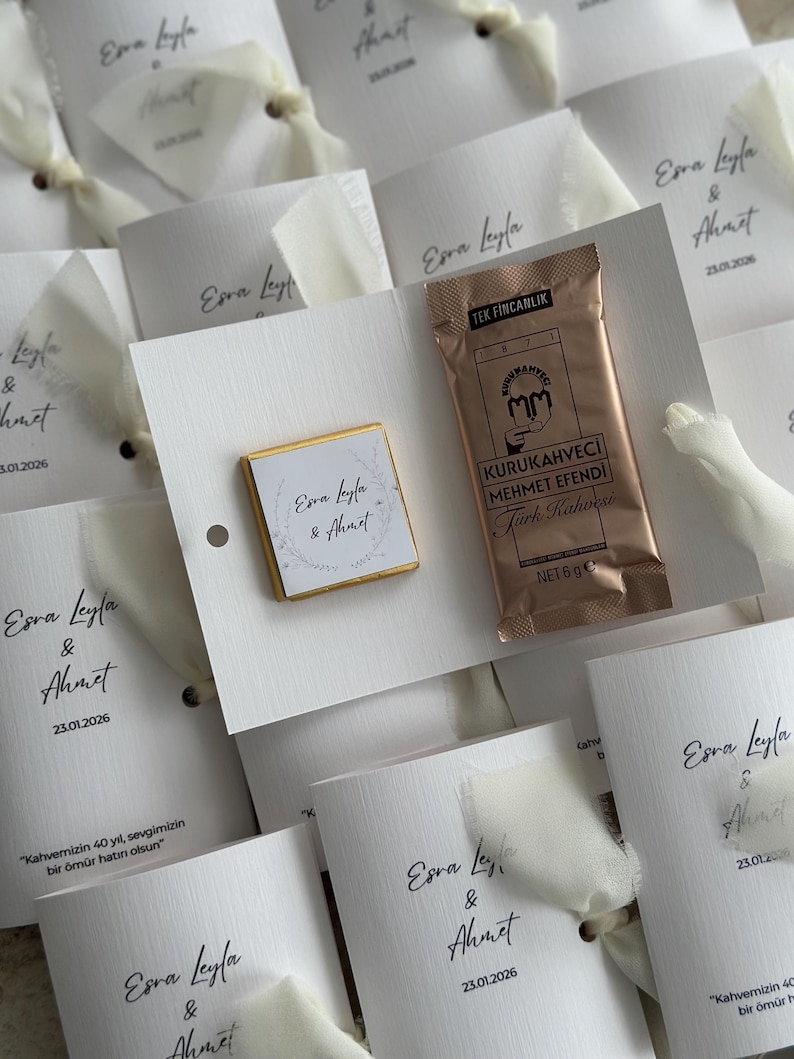 May include: A white card favor featuring a square chocolate with a floral wreath design and the names Esra Leyla & Ahmet, alongside a 6g packet of Kurukahveci Mehmet Efendi Turkish coffee, tied with a white ribbon and printed with the date 23.01.2026.