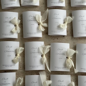 May include: A collection of white rectangular cards with a textured finish, each featuring the names Donda & Sebahattin, a date, and a short phrase in black script. Each card is adorned with a sheer, off-white ribbon tied in a bow through a hole.
