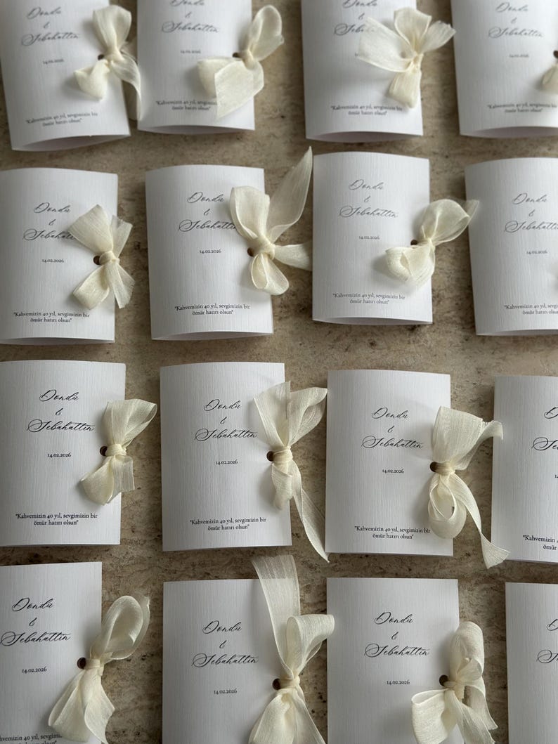 May include: A collection of white rectangular cards with a textured finish, each featuring the names Donda & Sebahattin, a date, and a short phrase in black script. Each card is adorned with a sheer, off-white ribbon tied in a bow through a hole.