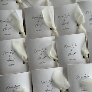 May include: A collection of white rectangular paper cards featuring the printed text Esra Leyla & Ahmet and the date 23.01.2026. Each card is accented with a cream-colored fabric ribbon tied through a small hole on the right side of the card.