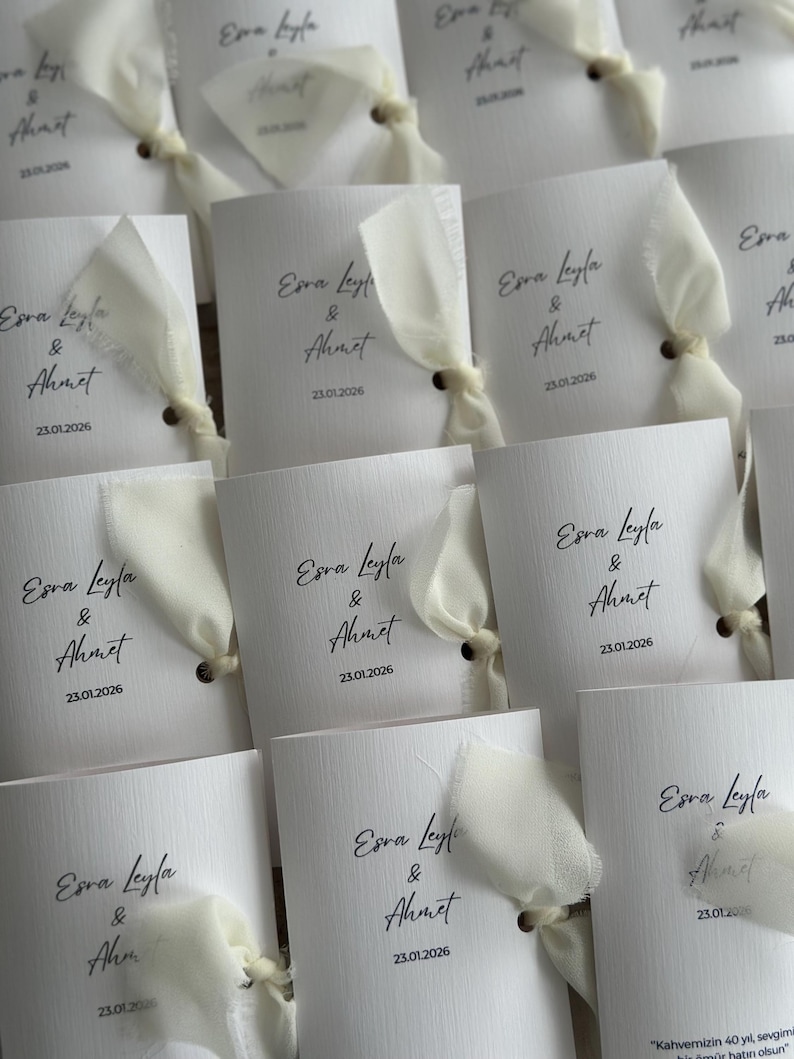 May include: A collection of white rectangular paper cards featuring the printed text Esra Leyla & Ahmet and the date 23.01.2026. Each card is accented with a cream-colored fabric ribbon tied through a small hole on the right side of the card.