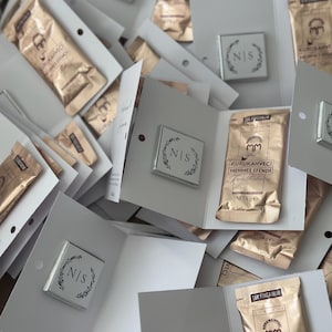 May include: A collection of white folded cardstock favors, each containing a square silver-toned emblem with the letters N|S surrounded by a leaf wreath and a gold-toned 6g packet of Kurukahveci Mehmet Efendi Turkish coffee with black text.