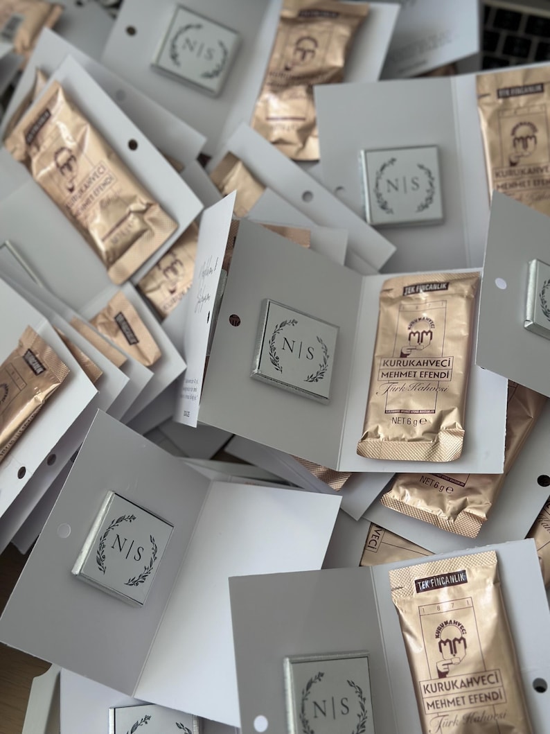 May include: A collection of white folded cardstock favors, each containing a square silver-toned emblem with the letters N|S surrounded by a leaf wreath and a gold-toned 6g packet of Kurukahveci Mehmet Efendi Turkish coffee with black text.