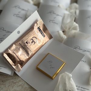 May include: A white cardstock folder containing a 6g packet of Kurukahveci Mehmet Efendi Turkish coffee and a square chocolate piece wrapped in gold foil with the initials R&E printed on a white label, surrounded by similar folded cards.
