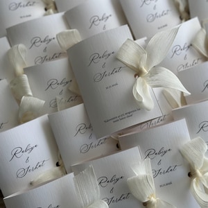 May include: A collection of white folded cards featuring the names Rabiya & Serhat in black cursive script, the date 21.11.2026, and the text "Kahvemizin 40 yıl, sevgimizin bir &ouml;m&uuml;r hatırı olsun". Each card is tied with a cream-colored fabric ribbon.