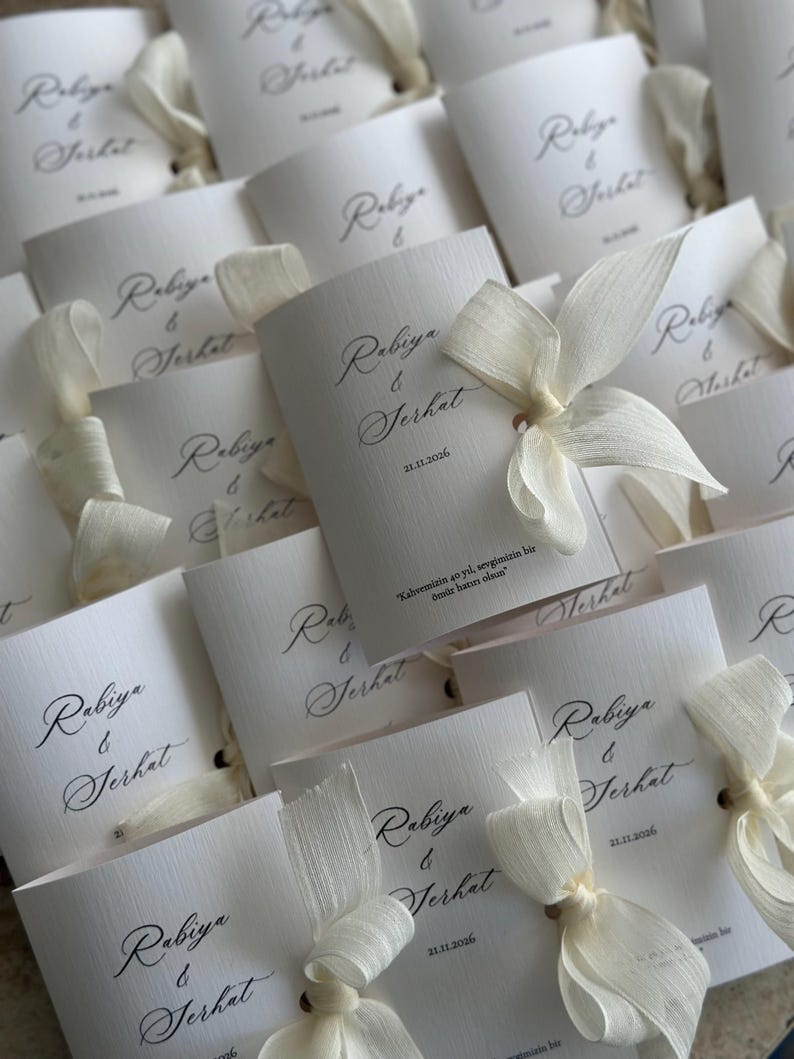 May include: A collection of white folded cards featuring the names Rabiya & Serhat in black cursive script, the date 21.11.2026, and the text "Kahvemizin 40 yıl, sevgimizin bir &ouml;m&uuml;r hatırı olsun". Each card is tied with a cream-colored fabric ribbon.