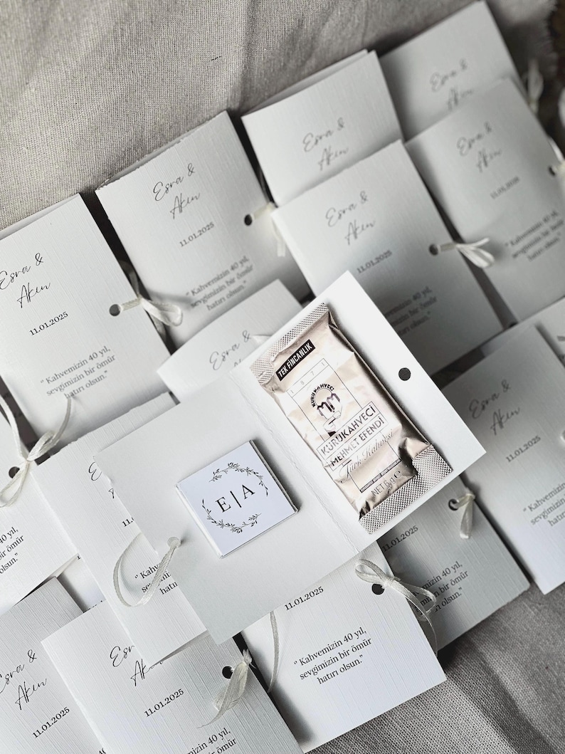 May include: A collection of white folded paper favor cards featuring the text Esra & Akin, the date 11.01.2025, and a Turkish quote. Each card contains a small square chocolate with an E|A monogram and a 6g packet of Kurukahveci Mehmet Efendi coffee.