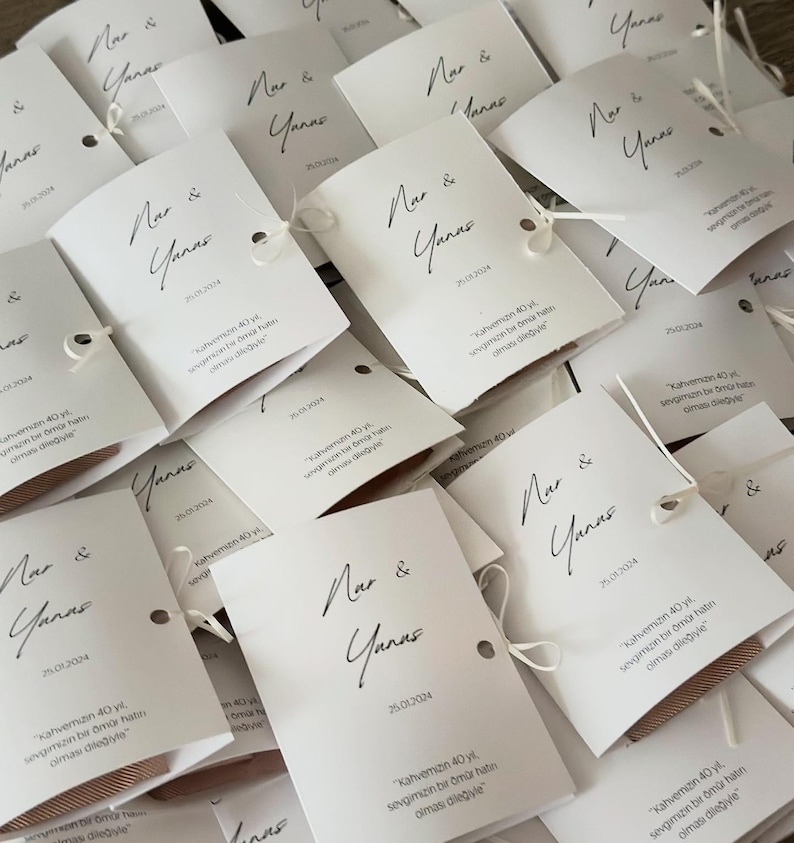 May include: A collection of white rectangular paper cards featuring the black printed names Nar & Yanas, the date 25.01.2024, and the Turkish text kahvemizin 40 yıl, sevgimizin bir &ouml;m&uuml;r hatırı olması dileğiyle. Each card has a small hole with white string.