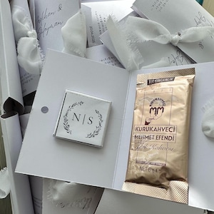 May include: A white cardstock gift set featuring a square chocolate bar with the initials N|S inside a botanical wreath and a gold-colored 6g packet of Kurukahveci Mehmet Efendi Turkish coffee, tied with a white frayed-edge ribbon.