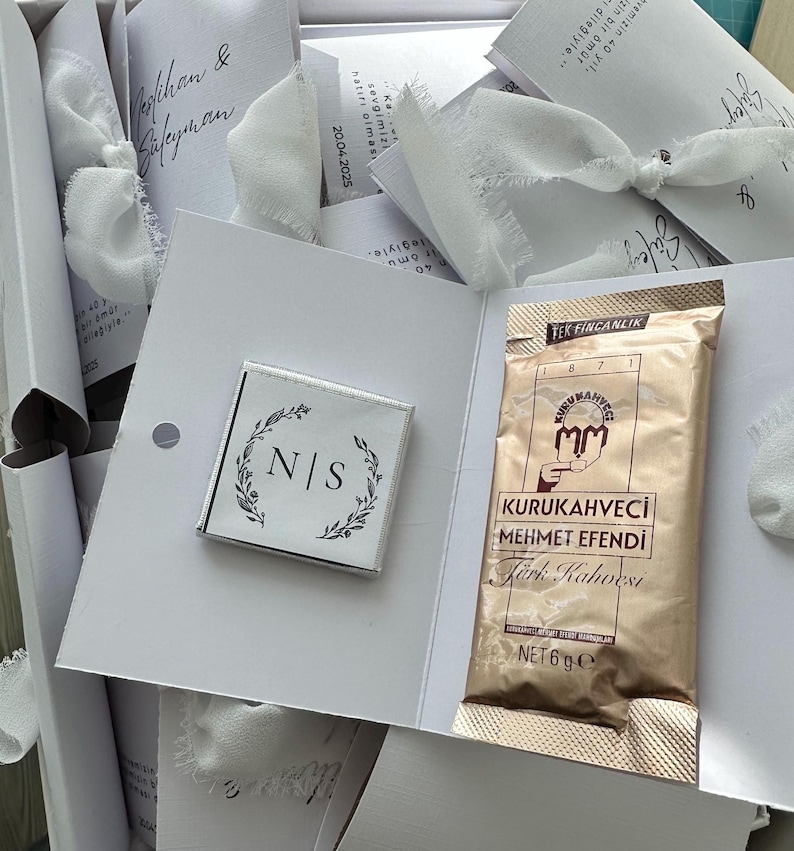 May include: A white cardstock gift set featuring a square chocolate bar with the initials N|S inside a botanical wreath and a gold-colored 6g packet of Kurukahveci Mehmet Efendi Turkish coffee, tied with a white frayed-edge ribbon.