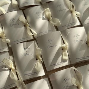 May include: A collection of white rectangular folded cards with a textured surface. Each card features the printed names Nefise & Jonmez in a dark script font, tied with a cream-colored ribbon, and includes the text Kahvemizin 40 yıl, sevgimizin bir &ouml;m&uuml;r hatırı olsun.