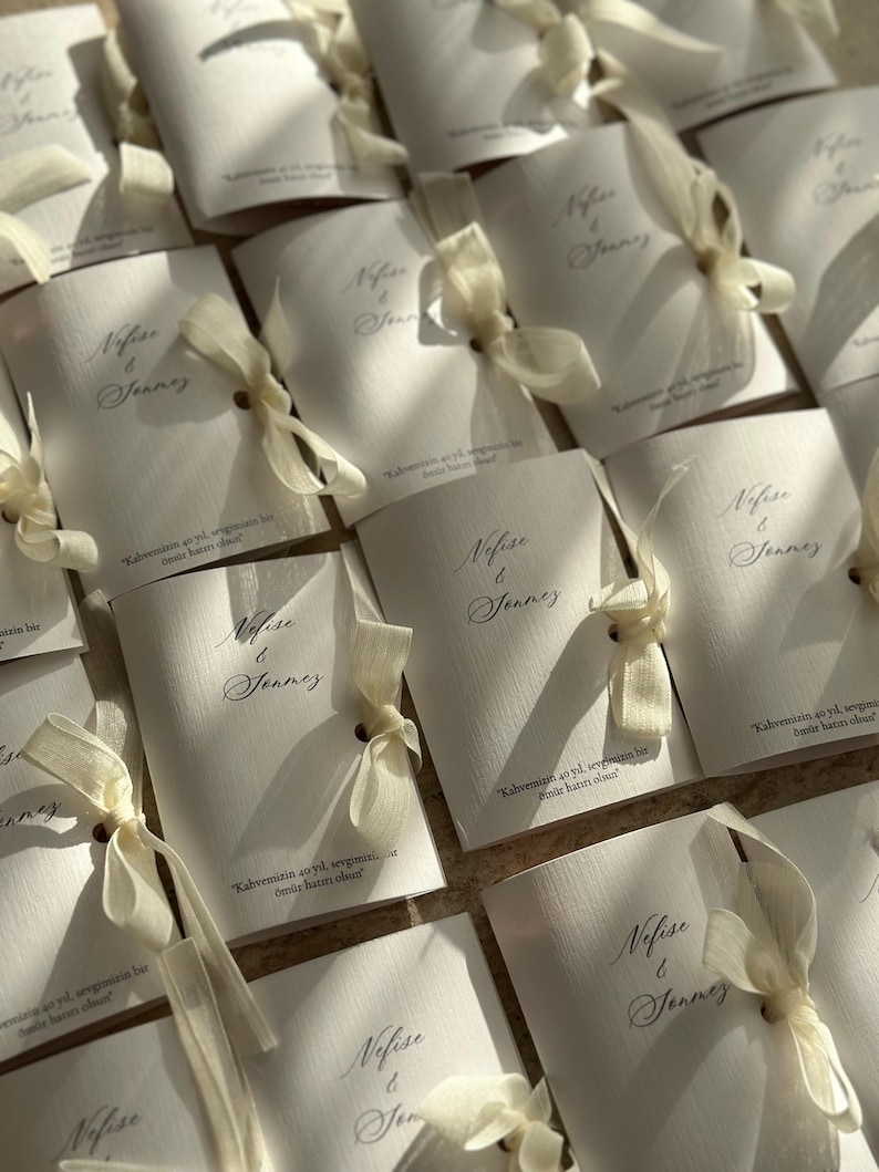 May include: A collection of white rectangular folded cards with a textured surface. Each card features the printed names Nefise & Jonmez in a dark script font, tied with a cream-colored ribbon, and includes the text Kahvemizin 40 yıl, sevgimizin bir &ouml;m&uuml;r hatırı olsun.