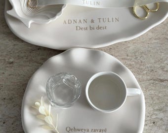 Puede incluir: Una bandeja ovalada de color blanco roto con un plato en forma de concha, tijeras doradas y cintas impresas con ADNAN y TULIN. Debajo, un plato redondo de color blanco roto contiene un vaso, una taza de cerámica blanca y el texto Qehweya zavayê Adnan.