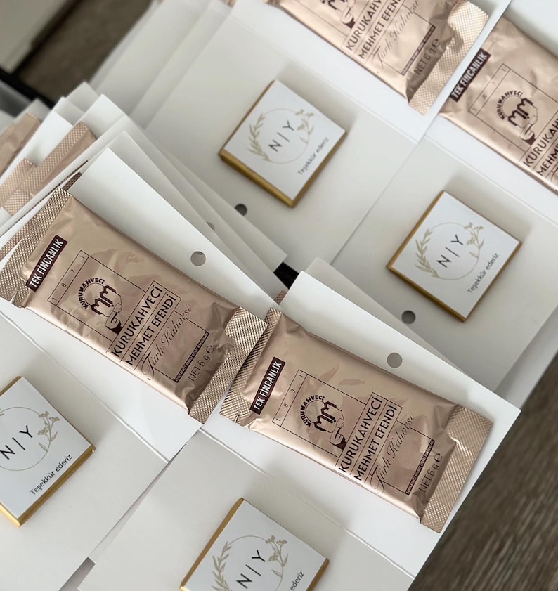 May include: A collection of rectangular Kurukahveci Mehmet Efendi Turkish coffee packets in metallic gold packaging, each labeled 6g, alongside small square white cards featuring a circular wreath graphic and the text NIY Teşekk&uuml;r ederiz.