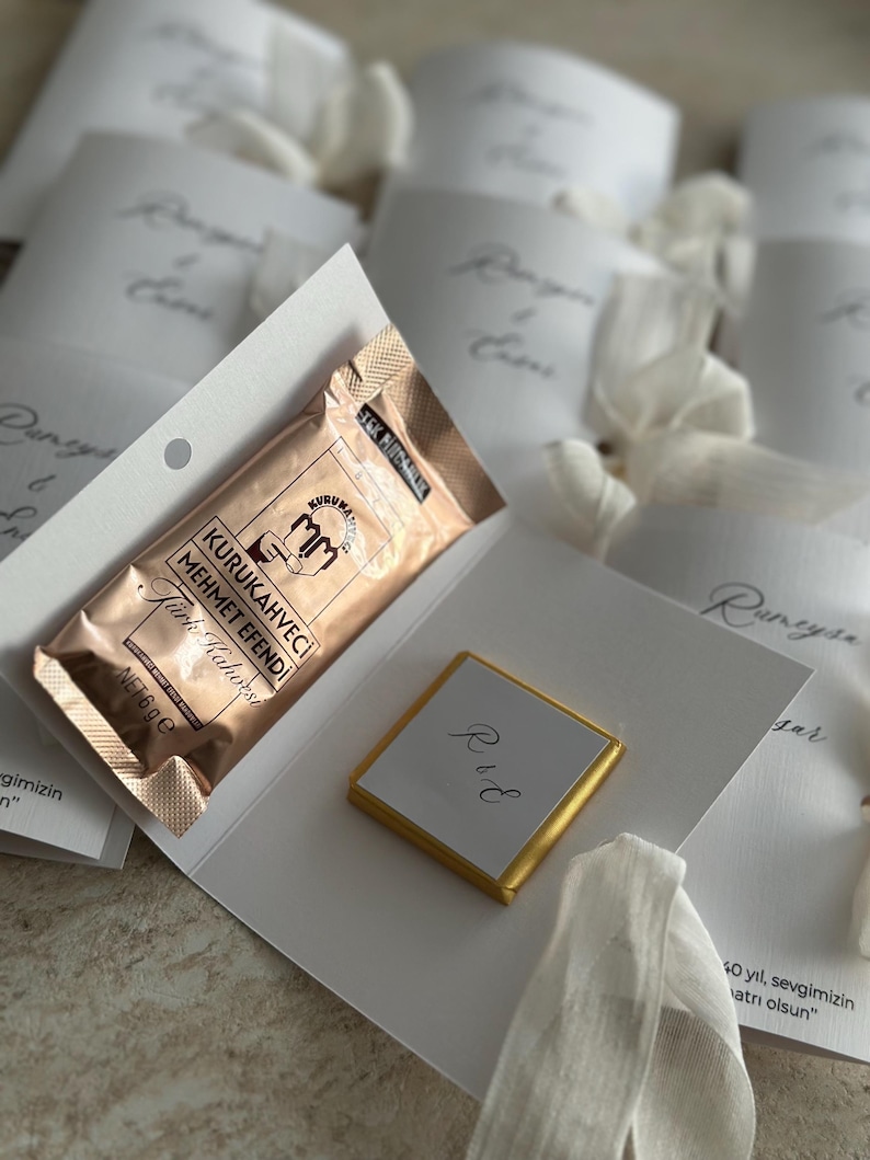 May include: A white cardstock folder containing a 6g packet of Kurukahveci Mehmet Efendi Turkish coffee and a square chocolate piece wrapped in gold foil with the initials R&E printed on a white label, surrounded by similar folded cards.
