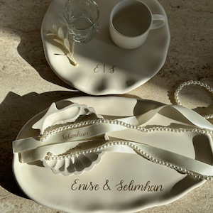 Op de afbeelding: Twee crèmekleurige keramische trays. Eén met een kopje, glas en decoratieve tak, met de initialen "E|S" in goud. De andere tray heeft de namen "Enise & Selimhan" en "Selimhan Enise" in gouden script, met een parelketting en lint.