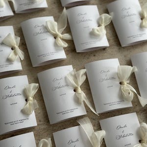 May include: A collection of white rectangular folded cards featuring the names Donda & Sebahattin, the date 14.02.2006, and Turkish text. Each card is accented with a sheer cream-colored ribbon tied in a bow through a small hole on the right side.