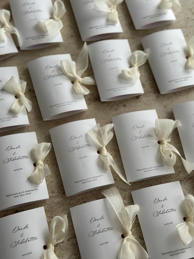 May include: A collection of white rectangular folded cards featuring the names Donda & Sebahattin, the date 14.02.2006, and Turkish text. Each card is accented with a sheer cream-colored ribbon tied in a bow through a small hole on the right side.