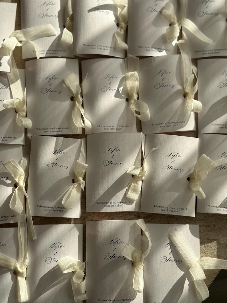 May include: A collection of white rectangular paper cards, each tied with a cream-colored ribbon. The cards feature the printed names Nefise & Sonmez in a cursive script, along with the text Kahvemizin 40 yıl, sevgimizin bir &ouml;m&uuml;r hatırı olsun.