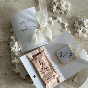 May include: A white card set tied with a cream ribbon, featuring the text Demet &ucirc; Umut Can. It includes a 6g packet of Kurukahveci Mehmet Efendi Turkish coffee and a small square chocolate with the letters D|U surrounded by leaf illustrations.