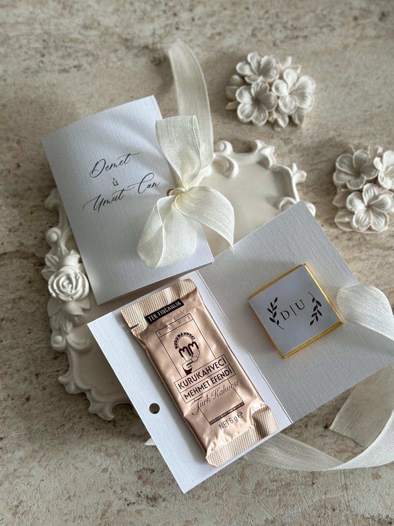 May include: A white card set tied with a cream ribbon, featuring the text Demet &ucirc; Umut Can. It includes a 6g packet of Kurukahveci Mehmet Efendi Turkish coffee and a small square chocolate with the letters D|U surrounded by leaf illustrations.