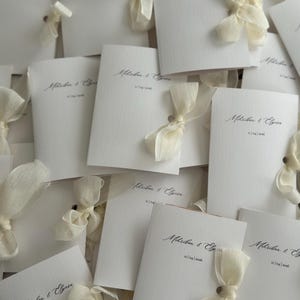 May include: A collection of white textured paper wedding invitations, each featuring the printed names Mihriban & Elyse and the date 11|04|2026, adorned with a delicate cream-colored sheer ribbon tied in a bow through a small hole in the card.