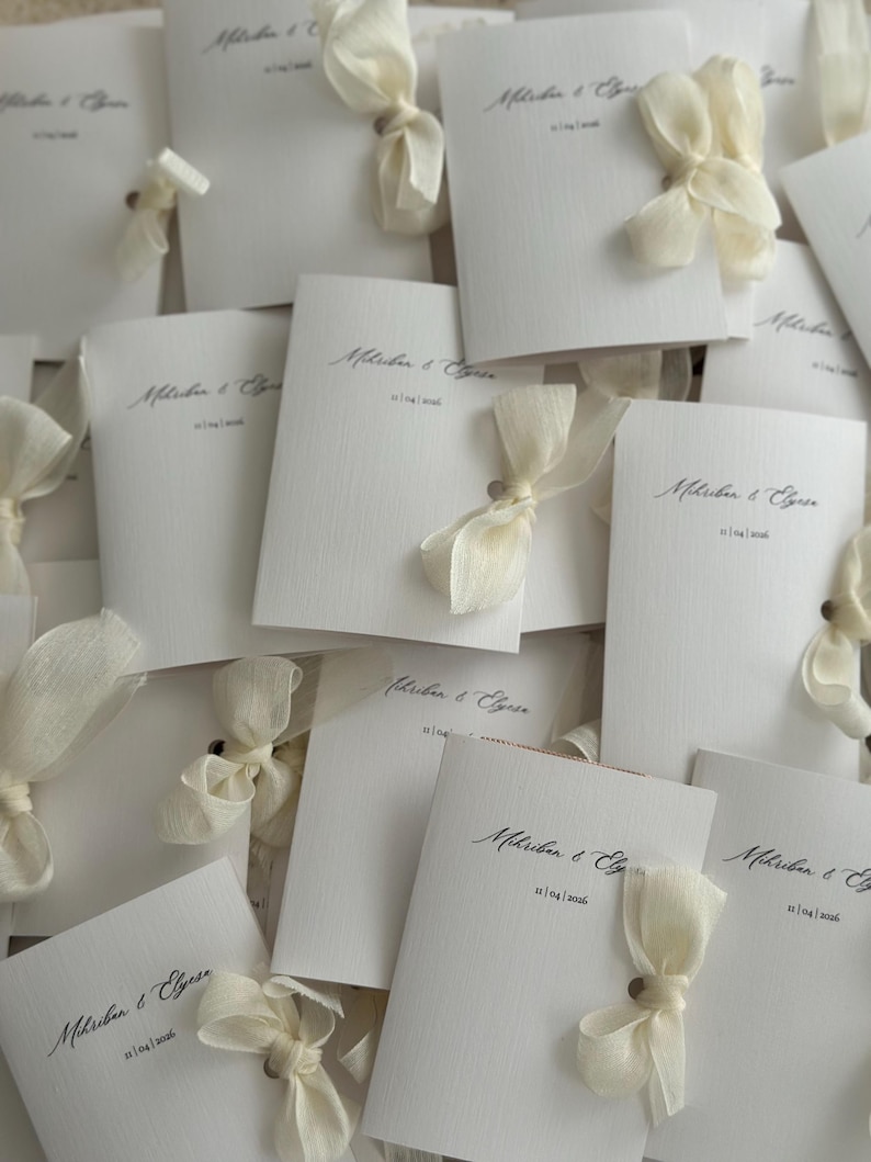 May include: A collection of white textured paper wedding invitations, each featuring the printed names Mihriban & Elyse and the date 11|04|2026, adorned with a delicate cream-colored sheer ribbon tied in a bow through a small hole in the card.