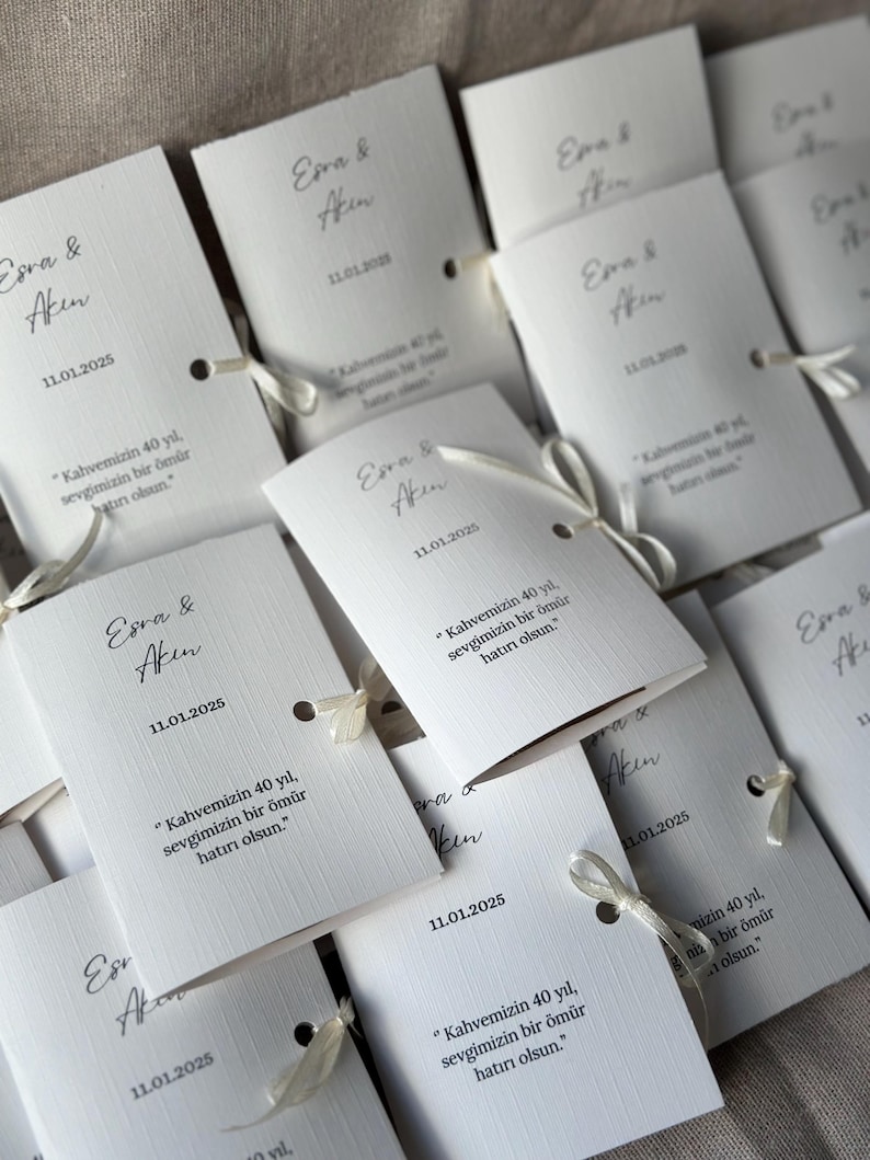 May include: A collection of white rectangular folded paper cards featuring the names Esra & Akin, the date 11.01.2025, and the Turkish text "Kahvemizin 40 yıl, sevgimizin bir &ouml;m&uuml;r hatırı olsun." Each card is secured with a thin white ribbon.