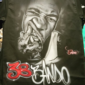 May include: Black and white portrait of a man with a gold grill, wearing a white tank top and a black shirt with the text "38 BANDO" in red and white spray paint.