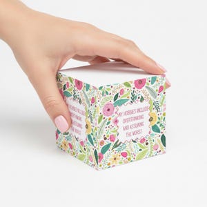 Peut inclure: Une boîte cadeau cubique blanche avec un motif floral rose, jaune et vert. Le texte "MY HOBBIES INCLUDE OVERTHINKING AND ASSUMING THE WORST" est imprimé sur la boîte. La boîte est tenue par une main.