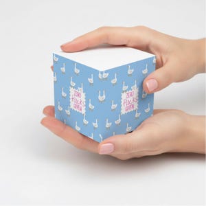 May include: A blue notepad cube with a pattern of white geese. The cube has the text "Zero flocks given" in a pink, postage stamp-like design. The notepad is held in two hands.