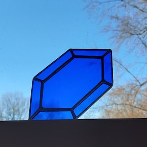 May include: A blue stained glass window hanging in front of a window. The glass is shaped like a diamond and has a dark blue border.