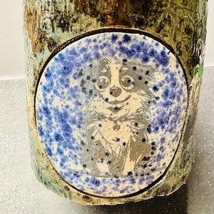 May include: A cylindrical ceramic vessel with a textured, mottled green and brown glaze. A circular inset panel features a white background with blue speckled paint and a black line-art illustration of a small, long-haired dog with large eyes.