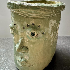 May include: A light green ceramic vessel shaped like a human head with a textured, uneven rim. The face features a prominent nose, a small mouth, and multiple dark-spotted eyes sculpted into the surface, finished with a glossy, mottled glaze.