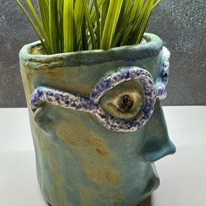 May include: A ceramic planter with a face design, featuring a blue and green color scheme. The planter has a detailed eye and glasses design. The planter is filled with green grass-like plants.