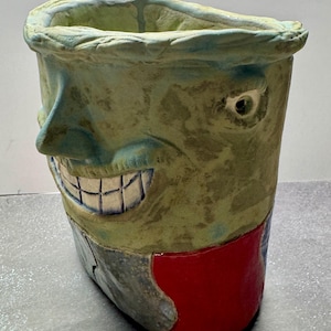 May include: A whimsical, handmade ceramic vase with a face design. The vase features a smiling face with blue and white teeth, a green and blue color scheme, and a red base. The unique, artistic design makes it a decorative piece.