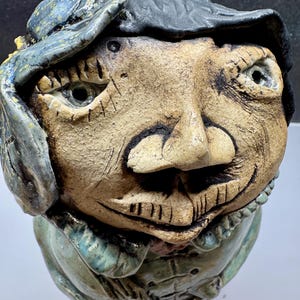 May include: A ceramic sculpture of a face with a hat. The face is tan with dark lines and details. The hat is blue and gray. The sculpture has a smiling expression and is wearing a blue jacket.