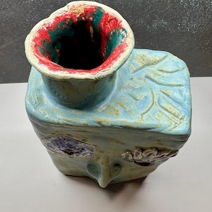 May include: A handmade ceramic owl-shaped vase with a light blue exterior and a red and green interior. The vase features textured details, including leaf patterns and facial features, and is approximately 6 inches tall.