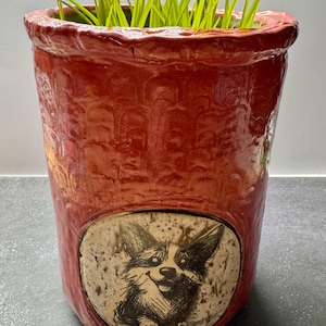 May include: A cylindrical red ceramic planter with a textured, scalloped surface pattern. A circular inset on the front features a detailed black and white sketch of a Corgi dog. The planter contains bright green grass blades extending from the top.