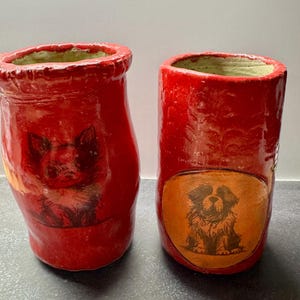 May include: Two cylindrical ceramic vessels with a glossy red glaze. The left vessel features a dark illustration of a cat, while the right vessel displays a dark illustration of a dog centered within a circular orange patch on the red surface.
