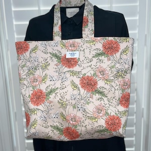 May include: A large tote bag with a floral pattern. The bag features a light pink background with a repeating pattern of orange and pink flowers, green leaves, and white accents. The handles and the top of the bag have the same floral pattern. The bag is displayed on a black shirt.