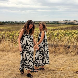 Puede incluir: Dos vestidos largos sin mangas con un estampado floral en blanco y negro. Los vestidos son usados por dos personas de pie en un campo de hierba seca y girasoles bajo un cielo nublado. Los vestidos tienen un corte holgado y se combinan con sandalias negras.