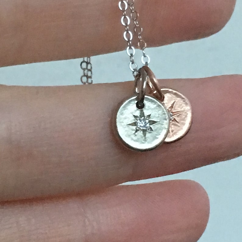 May include: A silver necklace with two charms. The larger charm is a silver circle with a small diamond in the center of a star design. The smaller charm is a rose gold circle with a star design.
