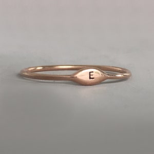 May include: A thin rose gold band ring with a small oval-shaped top that has the letter "E" engraved on it.