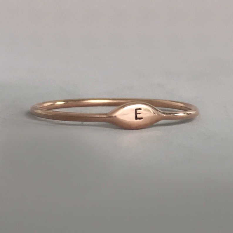 May include: A thin rose gold band ring with a small oval-shaped top that has the letter "E" engraved on it.