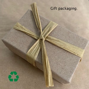 May include: A brown cardboard gift box tied with a beige ribbon. The box is wrapped in brown paper and tied with a beige ribbon. The ribbon is tied in a bow on top of the box. The box is sitting on a brown surface. Gift packaging.