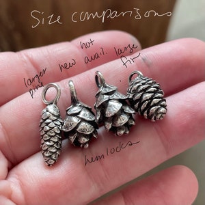 May include: A size comparison of four silver pine cone charms. The charms are arranged from largest to smallest. The largest charm is labeled "larger pine", the next largest is labeled "hemlocks", the next is labeled "new avail", and the smallest is labeled "large fir".