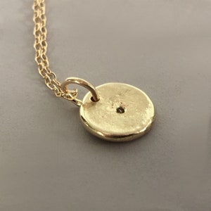 May include: A gold-colored pendant necklace featuring a small, round charm. The charm has a textured surface and a small hole near the top. The necklace chain is also gold-colored and delicate. The pendant is likely made of metal.