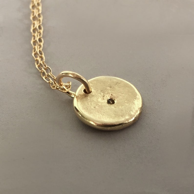 May include: A gold-colored pendant necklace featuring a small, round charm. The charm has a textured surface and a small hole near the top. The necklace chain is also gold-colored and delicate. The pendant is likely made of metal.