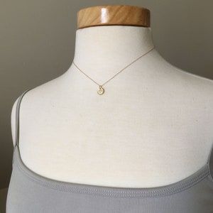 May include: A delicate gold necklace with a small, round pendant featuring a starburst design. The pendant is adorned with small, sparkling accents. The necklace is displayed on a mannequin wearing a gray tank top.